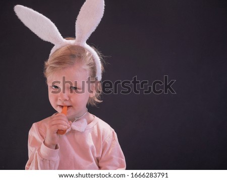 Funny blond child in pink pajamas and easter bunny costume eats carrot. Portrait of boy on black background