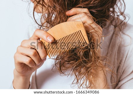 Young woman combing tangled ends of her curly hair with wooden comb while standing against white studio background, close-up.