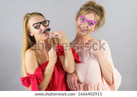 Happy two women holding paper decoration photo booth mask glasses and moustache on stick, having fun. Wedding, birthday and carnival funny accessories concept.