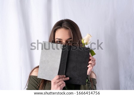 studio portrait of a girl. covered half of her face with a book with a dark cover. a white rose is seen from the book. concept art of intrigue and interest. fabric white fo
 Royalty-Free Stock Photo #1666414756