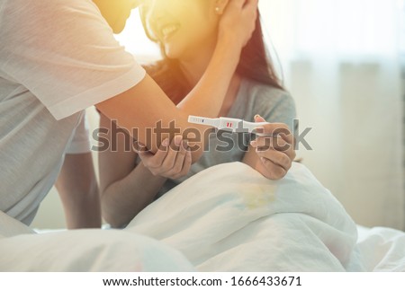 Cheerful and happy young asian man and woman with positive pregnancy test holding in hand while sitting on bed in bedroom at home. Royalty-Free Stock Photo #1666433671