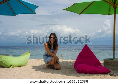 young beautiful and happy Asian Korean woman in chic Summer dress and sunglasses posing relaxed at tropical beach relaxed sitting at beanbag setup under umbrella parasol
