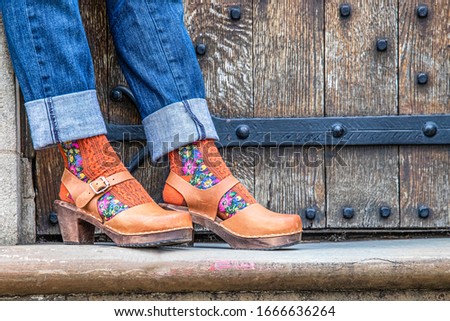 A beautiful woman wearing a pair of lovely brown leather and wood clog shoes standing in front of an olkd wooden vintage door Royalty-Free Stock Photo #1666636264