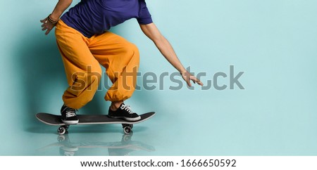 Bearded elderly man in t-shirt, orange pants and hat, gumshoes, bracelets. He crouched while riding black skateboard, posing sideways on blue background. Fashion, style, sport. Full length, copy space