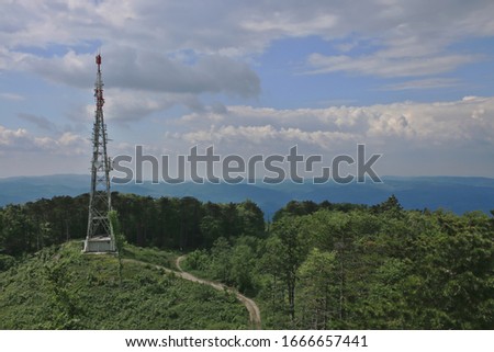 Cellular and broadcasting transmitter tower amidst a cleared spot of woodland full of grass, in the background wood, hill slopes, cloudy sky.