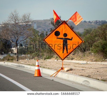 Orange diamond road sign with graphic of person holding a stop sign at a construction site. The sign has ornage flags on top and there is a sign reading Trucks Crossing in the distance
