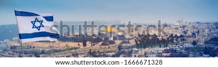 The holy land of Jerusalem with flag of Israel over the old city in haze. Cityscape of Jerusalem walls on the way of pilgrims and sacred place of three world religions - Christians, Muslims and Jews. Royalty-Free Stock Photo #1666671328