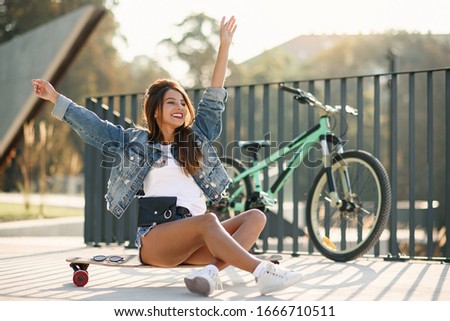Beautiful young cheerful woman sits on skateboard in urban space at sunrise.