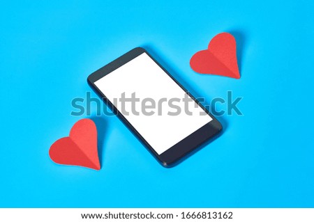 Black smartphone with isolated white screen for text, picture, photo and other graphics near two red paper hearts lies on blue table. Valentines day and love concept. Space for text. Close-up
