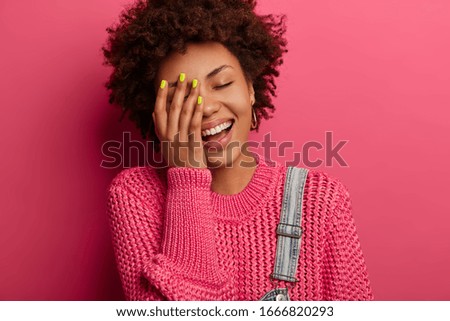 Carefree ethnic girl cannot stop laughing, keeps hand on face, has cheerful face, smiles positively, has good sense of humor, expresses happiness, wears knitted jumper, poses over pink background
