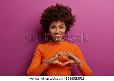 I love you with all my heart. Smiling dark skinned woman makes heart gesture, falls in love with someone, tells I am fully yours, has friendly look, dressed casually, isolated on purple background