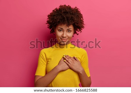 Sign of affection and admiration. Pretty curly woman presses palms to heart, being grateful for gift, wears yellow t shirt, poses over rosy background, says you are always in my heart, smiles gently Royalty-Free Stock Photo #1666825588