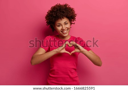 Pretty curly African American woman confesses in love, makes heart gesture, shows her true feelings, has happy expression, wears casual red t shirt, poses over pink background. Relationship concept