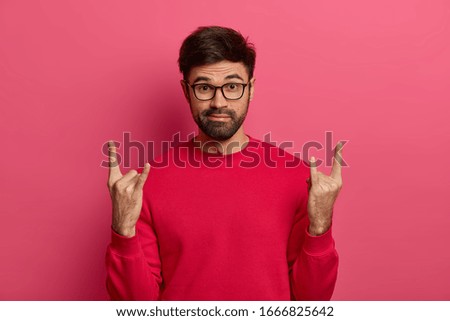 Amazed bearded guy visits awesome music festival, makes rock n roll gesture, has fun lisening favourite heavy meatal song, dressed casually, poses against pink background. Rock lives forever