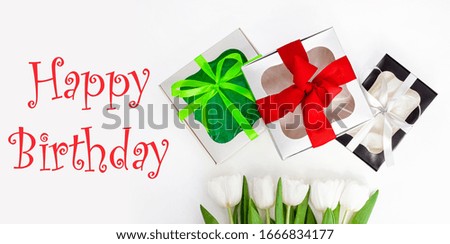 Happy Birthday text message sign on light background. White tulip flowers and paper gift boxes with colorful ribbon bows flat lay. Flower Bouquet congratulations greeting card. Website banner top view