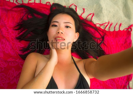 outdoors fresh and natural portrait of young beautiful and happy Asian Korean woman in bikini lying flat on sarong on beach sand taking selfie photo with mobile smiling cheerful and relaxed
