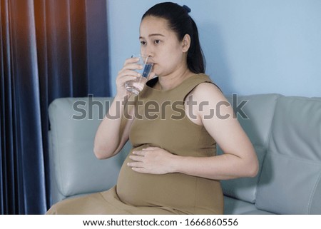 Young Asian pregnant woman holding  glass of water. Healthy living. Her sitting in liveing room. Nutrition and diet during pregnancy.