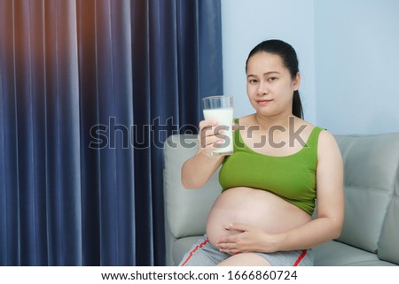 Young Asian pregnant woman holding  glass of milk. Healthy living. Her sitting in liveing room. Nutrition and diet during pregnancy.