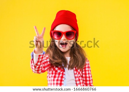 Cheerful stylish little girl dressed in pink checkered shirt, red cap, sunglasses, jeans on yellow background. Smiling cute child is showing with fingers peace sign, number two. Emotional portrait.