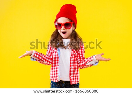 Beautiful cheerful stylish little girl dressed in pink checkered shirt, red cap, sunglasses and jeans on yellow background. Surprised shocked cute child is shrugging. Emotional portrait concept.