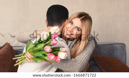 Happy holiday. Young lady with bouquet of fresh tulips hugging her beloved man at home, panorama