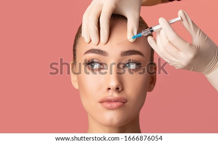 Skin Lifting Injection. Young Woman Receiving Botox Beauty Injection In Forehead Standing Over Pink Background. Studio Shot Royalty-Free Stock Photo #1666876054