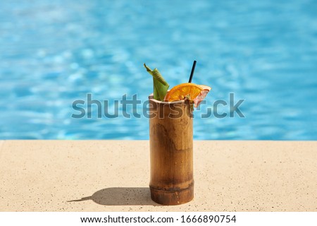 Outdoor picture of wooden cup with cocktail being near swimming pool, green and piece of orange being inside cocktail, black straw to drink, getting rid of thirst, cold drink in hot weather.