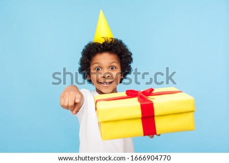 Funny excited joyful little boy with party cone on head pointing to camera and holding gift box, child choosing you, congratulating on birthday and giving awesome present. studio shot blue background