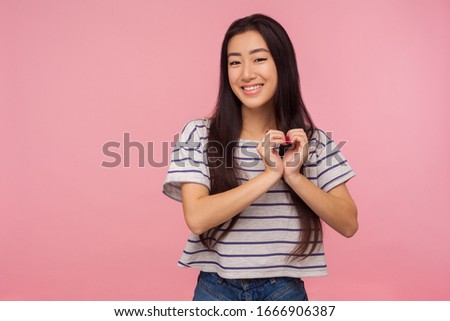 Portrait of charming lovely cute girl with long hair in striped t-shirt making heart shape with fingers and smiling, gesturing love, romantic feelings. indoor studio shot isolated on pink background