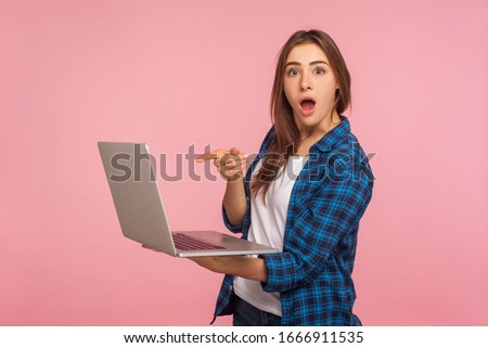 Portrait of shocked girl in casual checkered shirt pointing at laptop and looking at camera with amazement, surprised by computer software, application. studio shot isolated on pink background