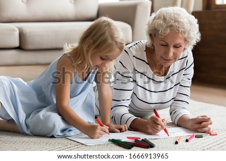 Happy loving mature grandmother lying on floor in living room drawing with cute little preschooler granddaughter, caring senior grandparent relax play have fun with small grandchild painting at home
