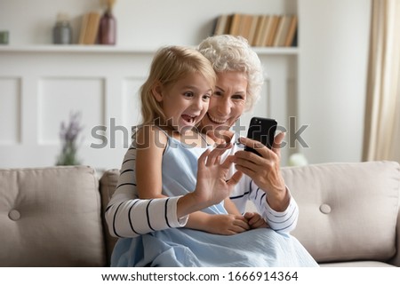 Overjoyed mature grandmother and cute little granddaughter sit on couch relax using smartphone together, happy senior granny and small grandchild have fun watch funny videos on modern cellphone