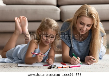 Happy young Caucasian mom or nanny lying on floor at home painting with cute little preschooler girl child, smiling mother relax rest with small daughter drawing together, early development concept