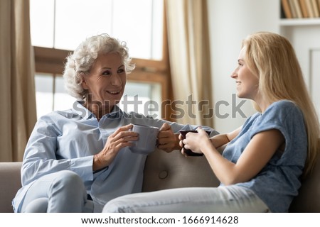 Smiling mature mother and adult daughter sit rest on sofa in living room chatting talking drinking tea or coffee, happy senior mom and grownup girl child relax on couch at home, enjoy time together Royalty-Free Stock Photo #1666914628