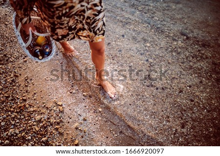 Cropped picture of female's legs walking in water. Woman holding picnic basket in hands.