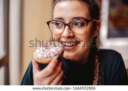 Portrait of smiling teenage girl sitting at pastry shop, holding doughnut and enjoying free time.