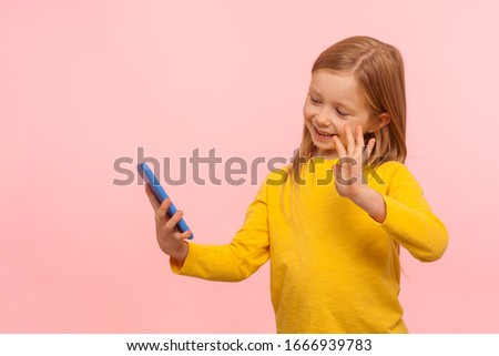 Portrait of happy beautiful little girl with ginger hair smiling and gesturing hi hello to mobile phone, child communicating with parents on video call. indoor studio shot isolated on pink background