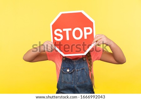 Prohibited way. Little girl in denim overalls covering face with octagonal Stop symbol, holding red traffic sign, attention to safe road crossing. indoor studio shot isolated on yellow background