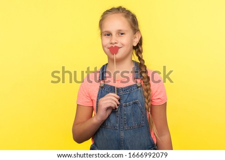 Portrait of charming happy little girl with braid in denim overalls covering mouth with red paper lips on stick and smiling at camera, symbol of kiss. indoor studio shot isolated on yellow background