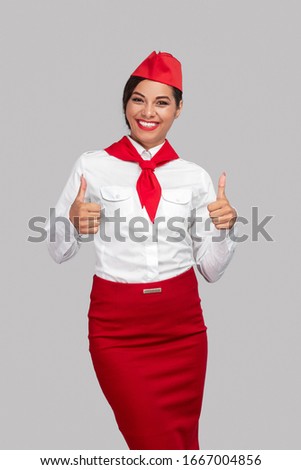 Joyful stewardess in bright red and white uniform smiling at camera and giving like while standing against gray background in studio