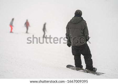 Unidentified tourist does snowboard skiing in Flachau, the ski resort in Austria. Man in black and grey sportswear. Three people on bokeh background. Foggy weather.