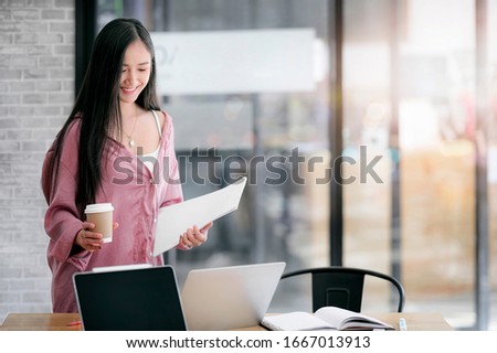 Young attractive asian woman holding cup of coffee and paperwork, looking at laptop scree and smiling while standing at office desk.