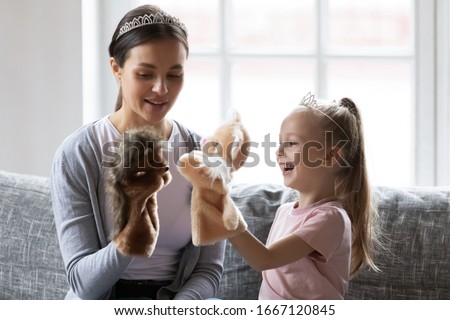 Head shot happy little girl wearing crown playing hand toys with pleasant young babysitter at home. Smiling mother enjoying daycare time with playful small child daughter, sitting together on couch. Royalty-Free Stock Photo #1667120845