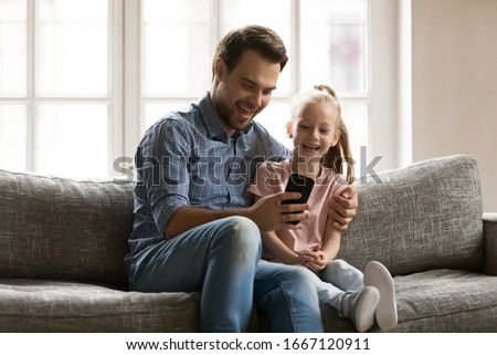 Laughing young daddy embracing small school aged child daughter, showing funny videos on smartphone. Overjoyed little girl looking at mobile screen with smiling father, technology addiction.