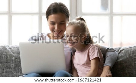 Head shot young smiling mother relaxing on comfy sofa with small school aged daughter, watching funny videos on laptop. Attractive happy nanny babysitter showing educational application to kid girl.