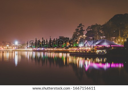 Beautiful long exposure landscape night picture of Xuan Huong lake, Da Lat, Vietnam at night. Neon lights of the buildings and their reflections create vibrant color trails on the water surface