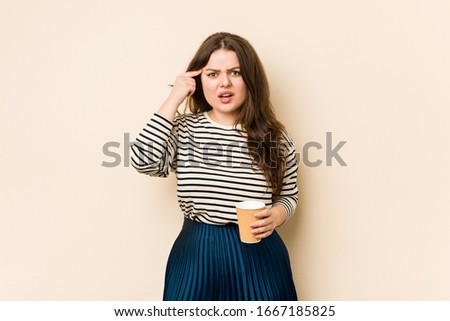 Young curvy woman holding a coffee showing a disappointment gesture with forefinger.