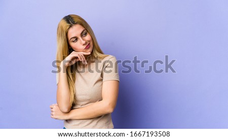 Young blonde woman isolated on purple background looking sideways with doubtful and skeptical expression.