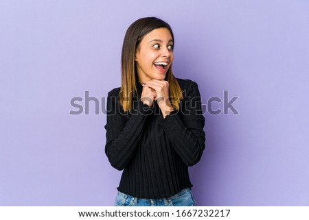 Young woman isolated on purple background praying for luck, amazed and opening mouth looking to front.