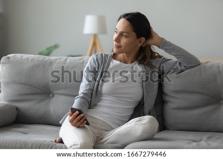 Smiling millennial Caucasian girl relax on cozy couch in living room hold modern smartphone thinking or pondering, happy young woman look in distance rest on comfortable sofa at home browsing cell